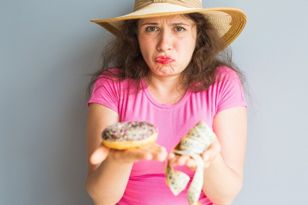 Confused young woman holding a donut and a measuring tape. Concept of Sweets, Unhealthy Junk Food and obesity.