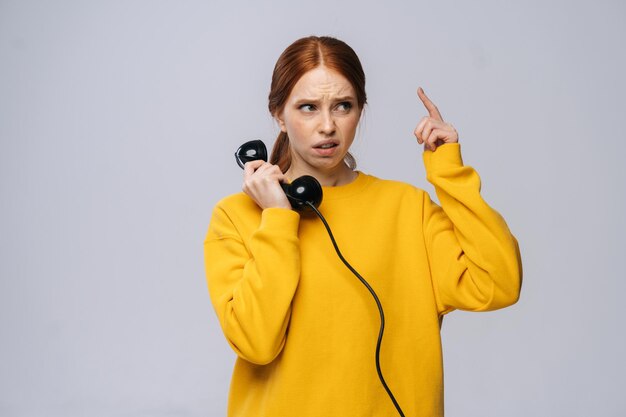 Confused young woman in stylish yellow sweater talking on retro phone and looking away