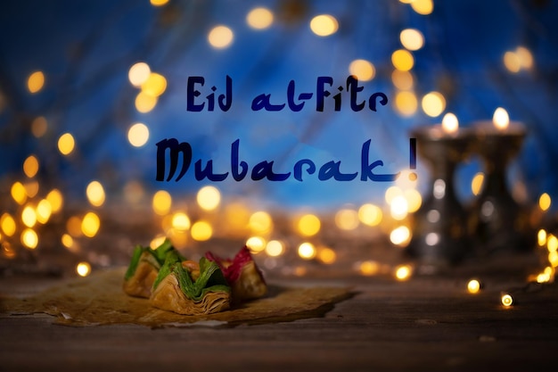 Congratulation eid alfitr mubarak arabic sweets on a wooden\
surface candle holders night light and night blue sky with crescent\
moon in the background