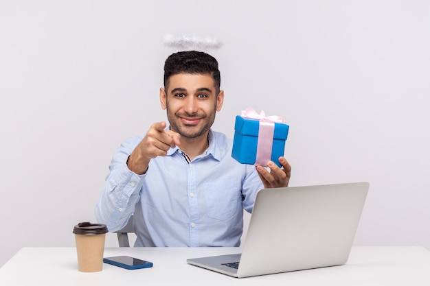 Congratulazioni a te gioioso uomo d'affari angelico con nimbo sulla testa che tiene una confezione regalo e indica la fotocamera seduta sul posto di lavoro del laptop saluto con vacanze professionali studio girato isolato