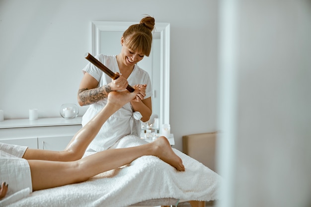 Уверенная женщина-специалист делает массаж ног бамбуковой палкой клиентке красивой кавказской женщины