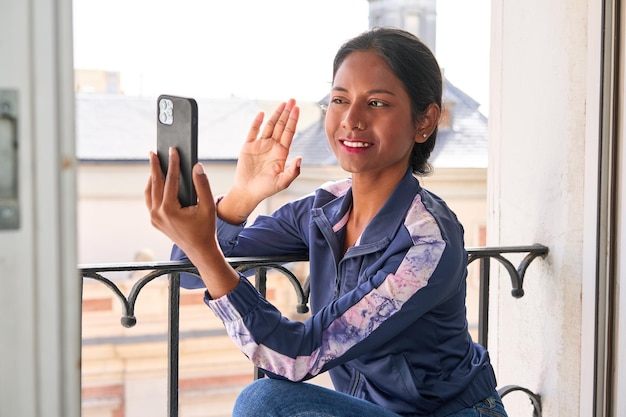 Подключенная и продуктивная молодая индийская женщина многозадачна по телефону, сидя на подоконнике
