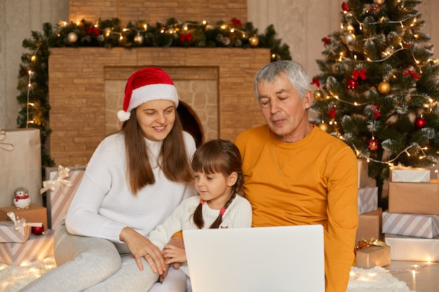 Connecting with family over video call during Christmas Eve, mother with little daughter and grandfather sitting on floor near xmas tree and looking at laptop screen.