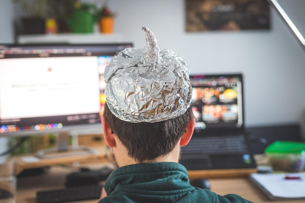 Conspiracy theory concept jonge man draagt een aluminium hoofd en zit achter de pc video's te kijken