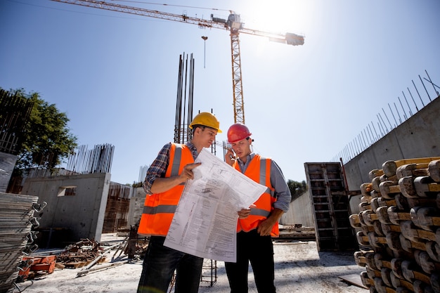 Constructeur en bouwmanager in oranje werkvesten en harde helmen bespreken documentatie en praten telefonisch op de open lucht bouwplaats naast de kraan. .