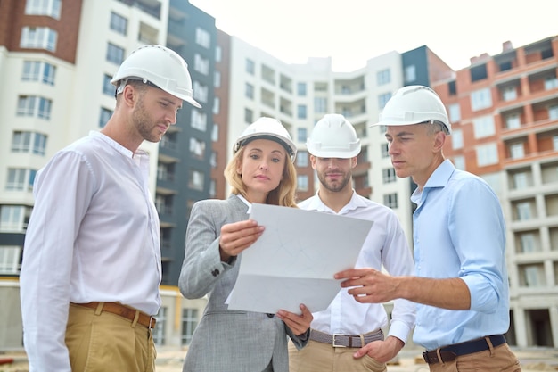Constructie plan. Zelfverzekerde vrouw in pak en veiligheidshelm en groep mannen in witte overhemden die 's middags naar tekening kijken terwijl ze op de bouwplaats staan