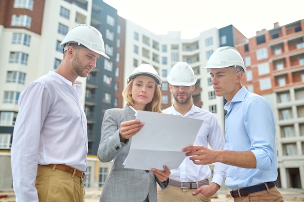 Constructie plan. zelfverzekerde vrouw in pak en veiligheidshelm en groep mannen in witte overhemden die 's middags naar tekening kijken terwijl ze op de bouwplaats staan