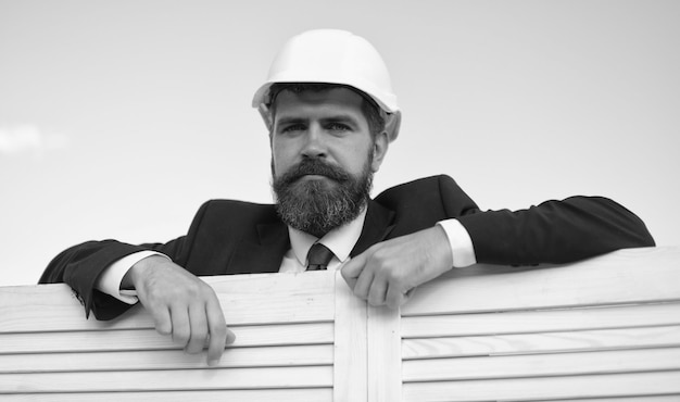 Construction and architecture concept Man with beard and happy face wears white helmet Foreman leans on wooden fence close up Architect in formal suit and hardhat on blue sky background