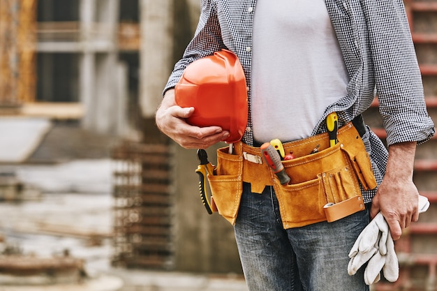 Immagine ritagliata per la sicurezza della costruzione di un costruttore maschio con casco e cintura degli attrezzi che lavora alla costruzione