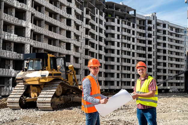На строительной площадке один рабочий в униформе показывает другому рабочему в униформе план дома Концепция строительства