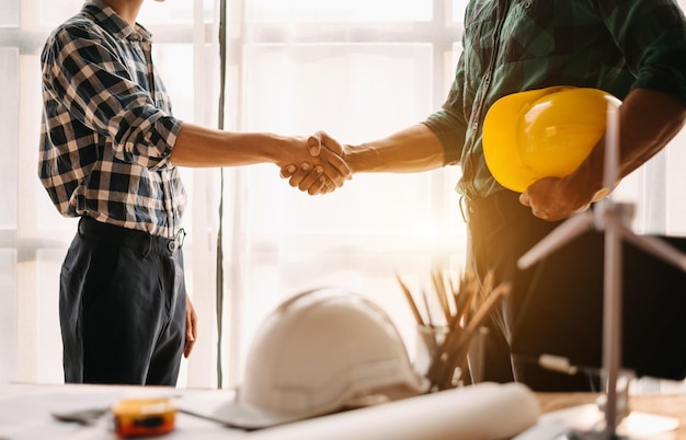 Construction team shake hands greeting start new project plan behind yellow helmet on desk in office center to consults about their building project in office