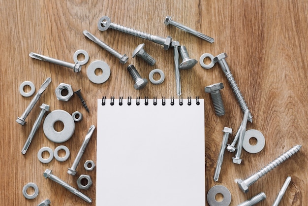 Construction tools. The screws, nuts and bolts arranged around blank spiral bound note book paper on wooden background. Repair, home improvement concept. Free space for text, top view, flat lay.