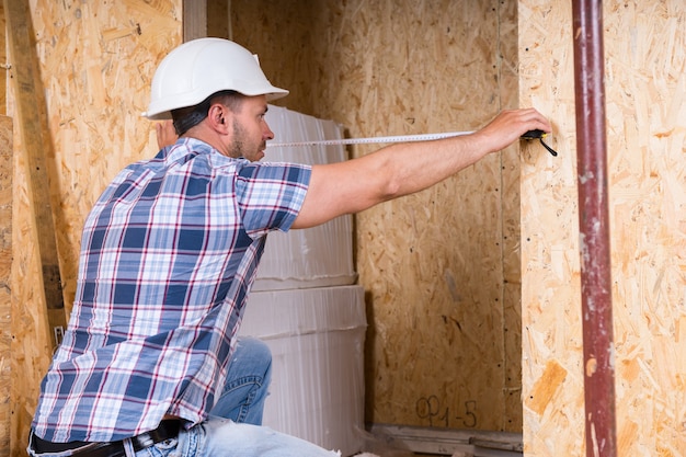 Construction Worker Builder Wearing White Hard Hat Measuring Width of Door Frame with Measuring Tape in Unfinished Home