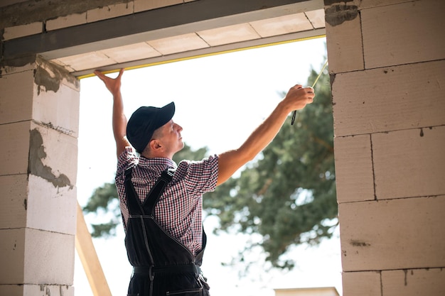 Construction worker at construction site measures the length of window opening and brick wall with tape measure Cottage are made of porous concrete blocks work clothes jumpsuit and baseball cap