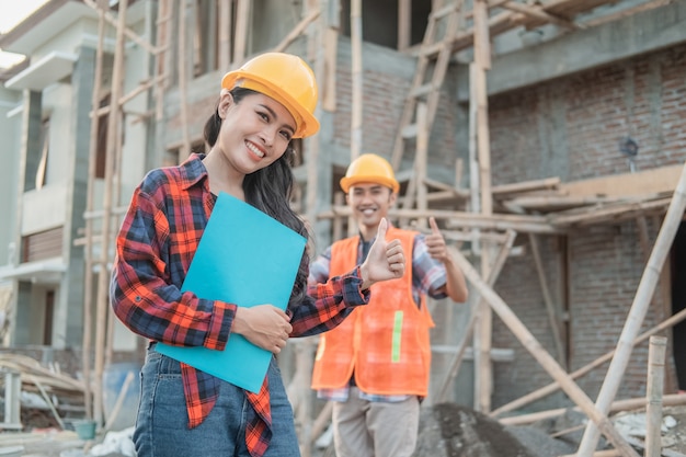 construction worker smiling at the camera wearing a safety helmet with a thumbs up against the background of an unfinished building