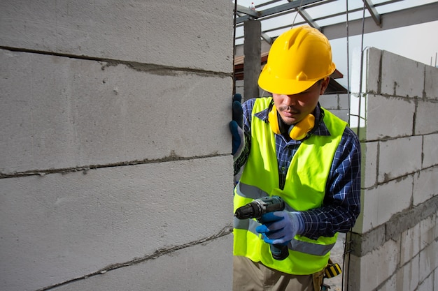 건설 노동자 사용 드릴 비트, 엔지니어 입고 안전 장비 헬멧 및 재킷 파워 드릴을 사용하여 화난 된 벽돌 벽.