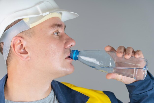 A construction worker in a white helmet and work clothes drinks water from a plastic bottle, close up foto