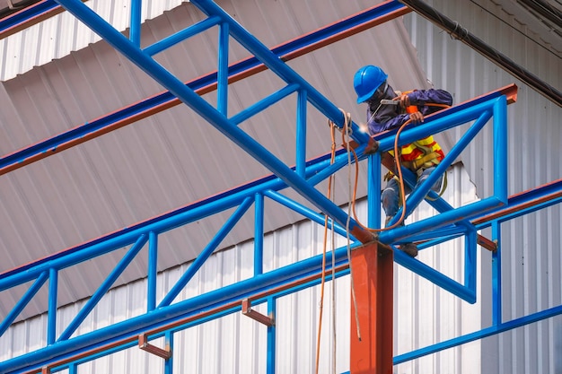 Construction worker with safety equipment is welding metal roof warehouse building structure