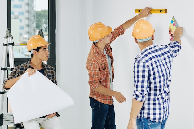 Construction workers discussing how to pain walls in empty room, their colleague checking blueprint