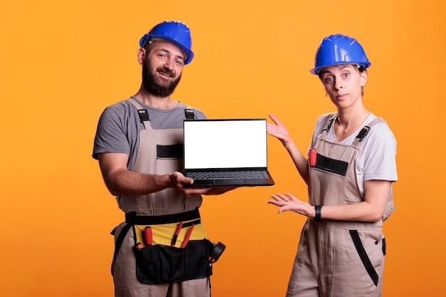 흰색 디스플레이가 있는 노트북을 들고 유니폼과 헬멧을 쓴 건설 노동자들. 휴대용 컴퓨터를 사용하여 모형 템플릿에 격리된 카피스페이스가 있는 빈 빈 화면을 표시합니다.