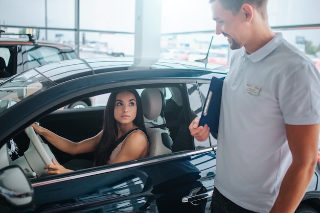 Consultant in white shirt stands besides car and looks at young woman