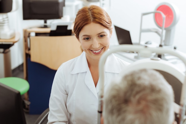 Consulting man. Beaming eye doctor wearing white coat sitting near laptop while consulting man