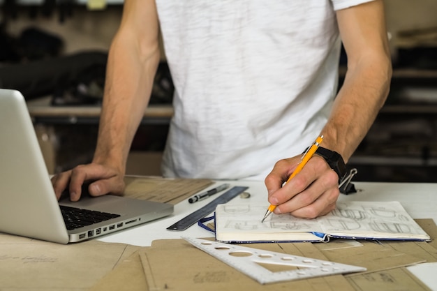 Consumentenaccessoires of tassenontwerper aan het werk in de werkplaats. Handen van jonge mannelijke persoon met potlood, ontwerpdocument en laptop computer bij een werktafel van kleine vervaardigingsfaciliteiten of bureau