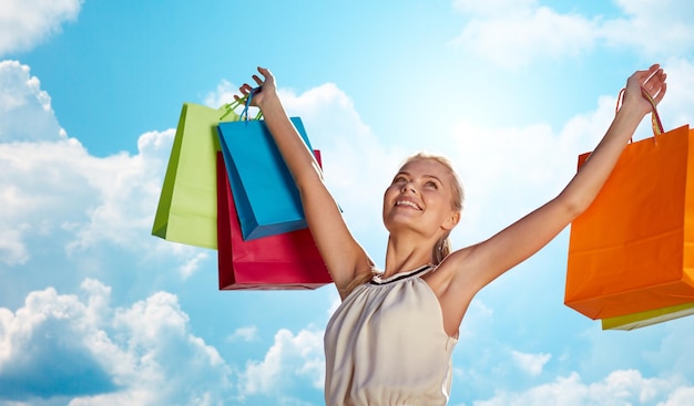 consumerism, sale and people concept - smiling woman with shopping bag rising hands over blue sky and clouds background