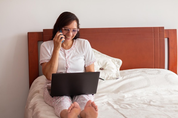 Contagious laughter a mature woman enjoys a phone conversation in bed while working on her laptop