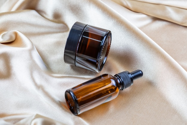 A container for storing cosmetics made of dark glass lie on the folds of a silk satin fabric. Perfume, serum or lotion bottle