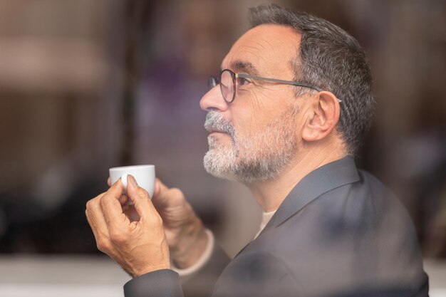 contemplative mature man with glasses looking out the window holding a small espresso