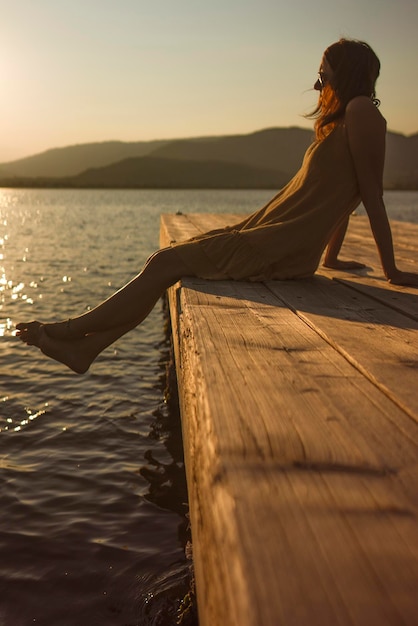 Una giovane donna caucasica contemplativa si siede da sola su un molo sul lago a fissare l'acqua al tramonto o all'alba in un'atmosfera arancione e vintage