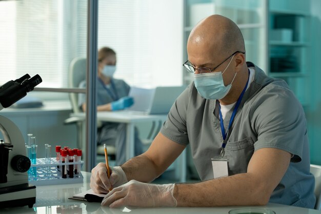 Contemporary bald male scientist in protective mask and gloves making scientific notes about new vaccine during research in laboratory