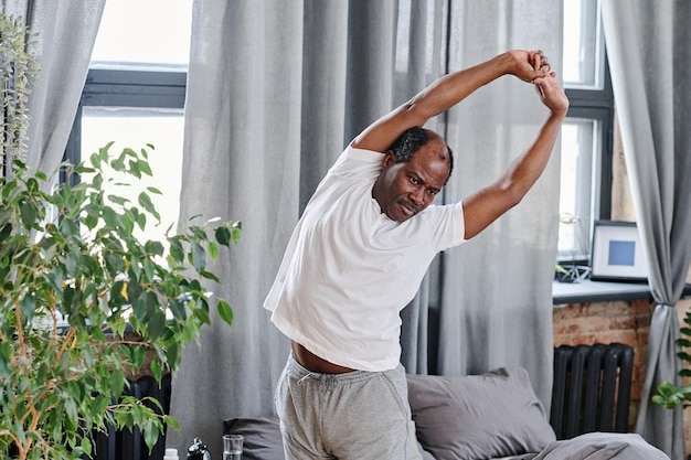 Contemporary elderly man in pajamas doing side bends by his bed after sleep