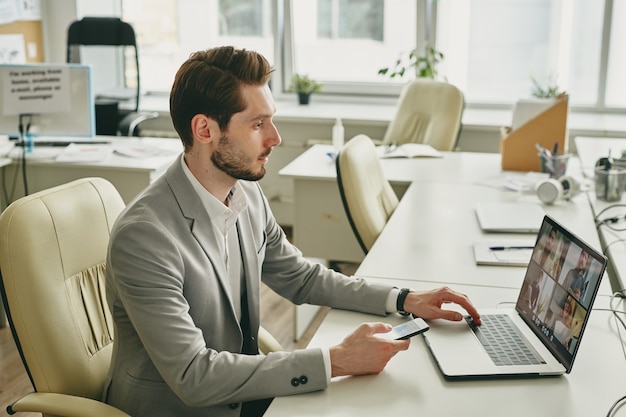 Contemporary elegant businessman looking at his colleagues working from home on laptop display while sitting in armchair by desk in office