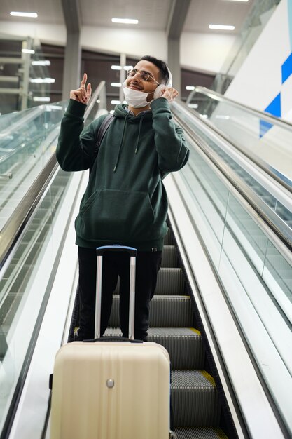 Contemporary happy young man in casualwear enjoying his favorite music in headphones while standing on step of moving escalator in airport