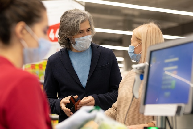 Contemporary mature husband and wife in protective masks looking at one another while deciding whether to pay by card or cash