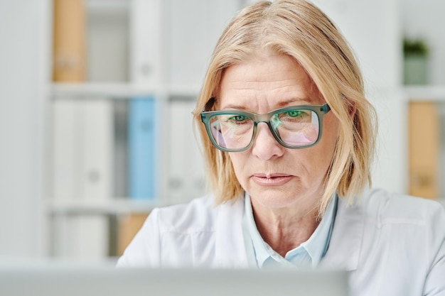 Contemporary mature online doctor looking at computer screen in office