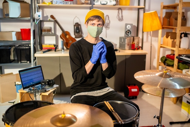 Contemporary musician in casualwear, protective gloves and mask sitting in front of drumset, making new music and recording it in garage