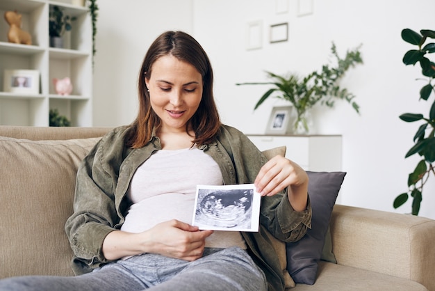 Donna incinta contemporanea che ti mostra l'immagine ultrasonica del futuro bambino e la guarda mentre ti rilassi nell'ambiente domestico