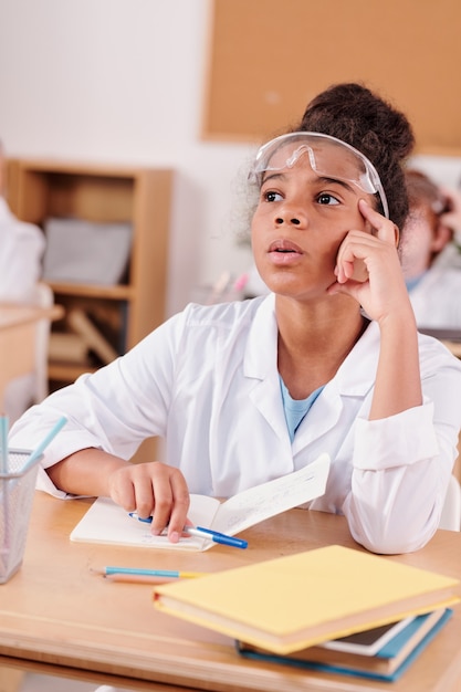 Contemporary schoolgirl of african ethnicity listening attentively at teacher of chemistry during experiment while sitting on classmates