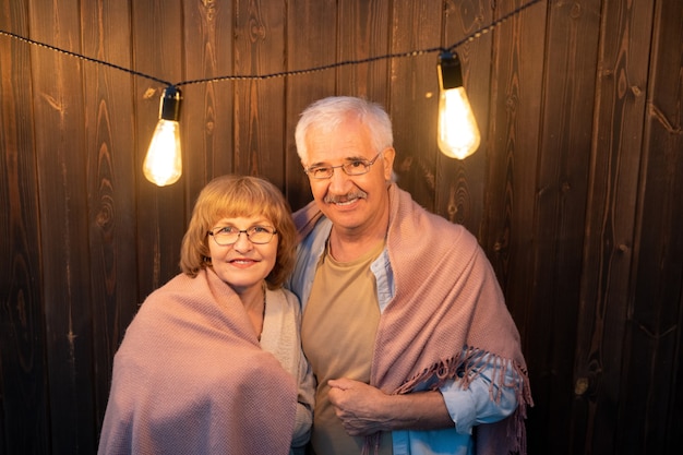 Contemporary senior couple wrapped into plaid standing against wall