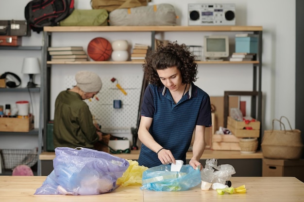 Contemporary teenage guy sorting waste in two large cellophane sacks