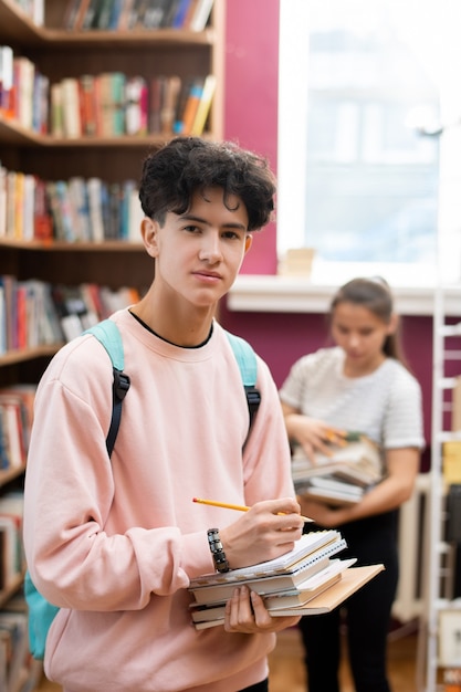 Contemporary teenager with backpack, stack of books and pencil standing in college library with classmate