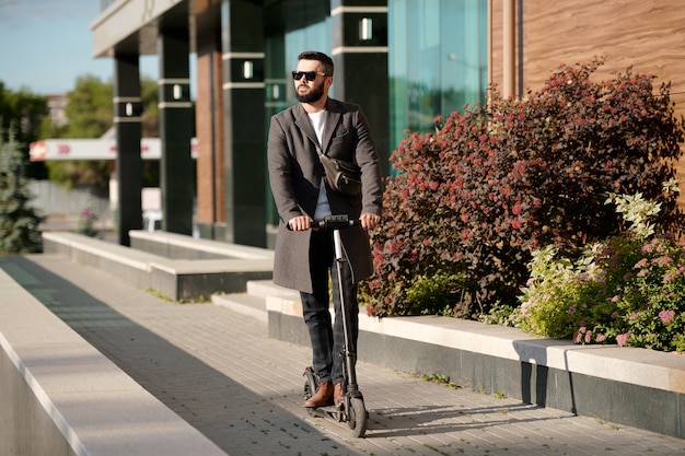 Contemporary young bearded businessman in coat and sunglasses riding on electric scooter in urban environment along plants and bushes