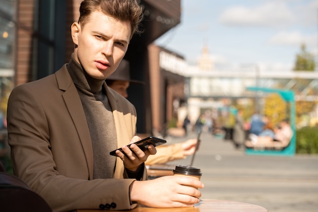 Contemporary young elegant man in jacket and sweater using smartphone while sitting by small table and having drink in urban environment