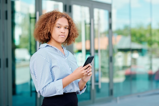 Contemporary young mobile employee with smartphone looking at you outdoors while texting