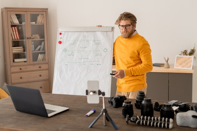 Contemporary young teacher with highlighter standing by whiteboard in front of laptop and smartphone camera during explanation