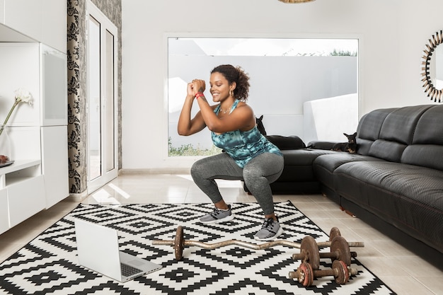 Content African American sportswoman doing squats while watching online video tutorial on laptop during home workout in living room with dogs