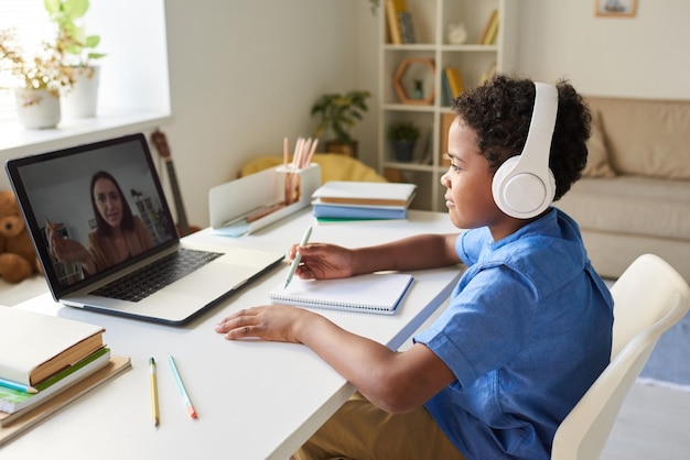 Photo content africanamerican schoolboy sitting at table and attentively listening to online tutor in head